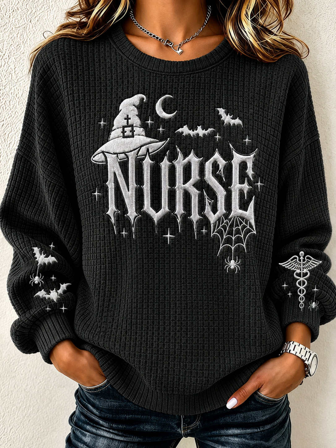 A woman wearing a black waffle-knit crewneck sweater with a gothic 'NURSE' graphic featuring a witch hat, bats, spiderwebs, and a caduceus symbol on the sleeves, paired with dark blue jeans.
