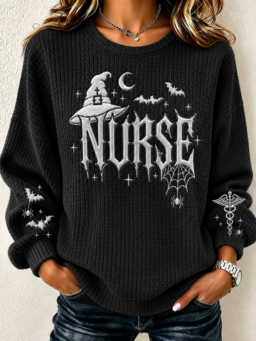 A woman wearing a black waffle-knit crewneck sweater with a gothic 'NURSE' graphic featuring a witch hat, bats, spiderwebs, and a caduceus symbol on the sleeves, paired with dark blue jeans.