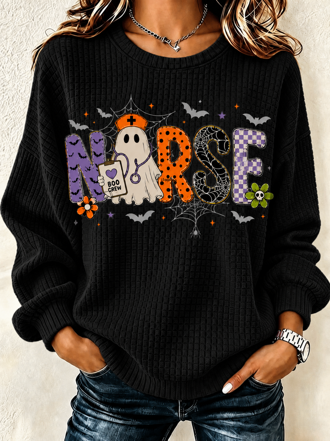 A woman wearing a black ribbed knit sweater with a colorful Halloween-themed 'Nurse' graphic, paired with blue jeans.