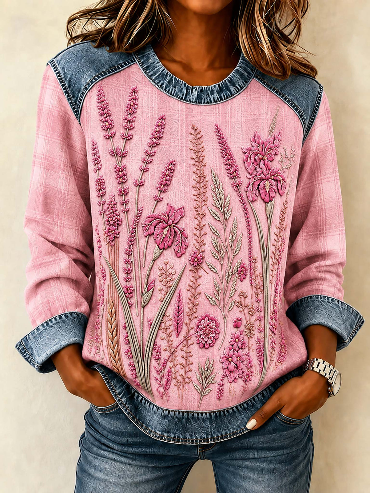 A woman wearing a pink long-sleeve top with blue denim shoulder panels and cuffs, featuring embroidered pink and green wildflower patterns on the front.