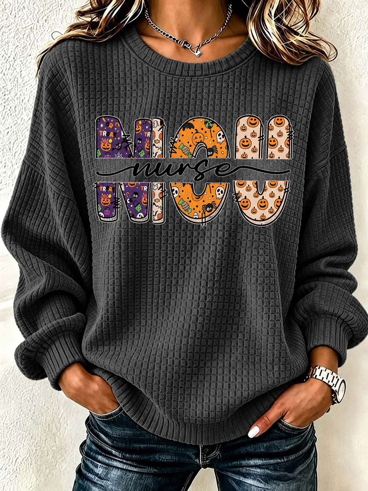 A woman wearing a dark gray waffle-knit crewneck sweatshirt with a 'MOON NURSE' graphic across the chest. The letters of 'MOON' feature Halloween-themed patterns including pumpkins, ghosts, and spiderwebs, while 'NURSE' is written in black script.