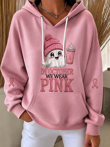 A person wearing a pink pullover hoodie with a graphic that reads 'IN OCTOBER WE WEAR PINK' and a cartoon ghost in a pink hat holding a pink drink. Pink awareness ribbons appear on both sleeves, and the person is also wearing white pants.