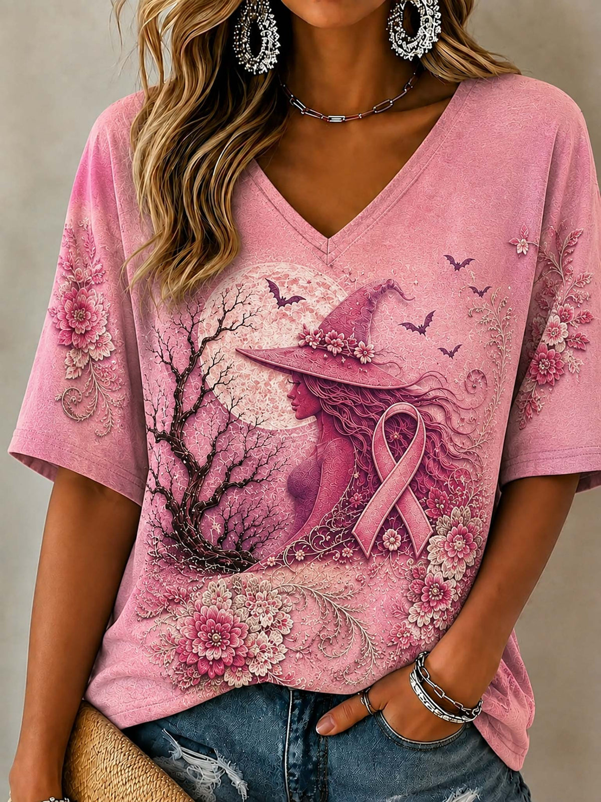 A woman wearing a pink V-neck short-sleeve t-shirt with a graphic print of a witch, a pink awareness ribbon, a full moon, and flowers. She is also wearing blue jeans, silver hoop earrings, and bracelets.