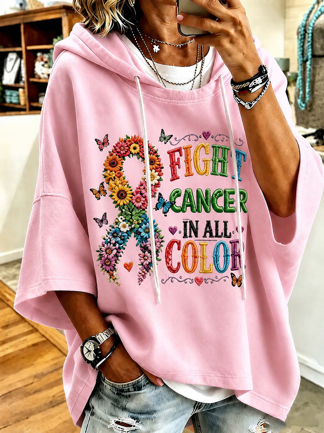A person wearing a light pink oversized hoodie with a colorful 'FIGHT CANCER IN ALL COLORS' graphic and a floral ribbon design, paired with distressed denim jeans in an indoor setting.