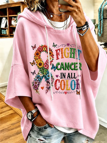 A person wearing a light pink oversized hoodie with a colorful 'FIGHT CANCER IN ALL COLORS' graphic and a floral ribbon design, paired with distressed denim jeans in an indoor setting.