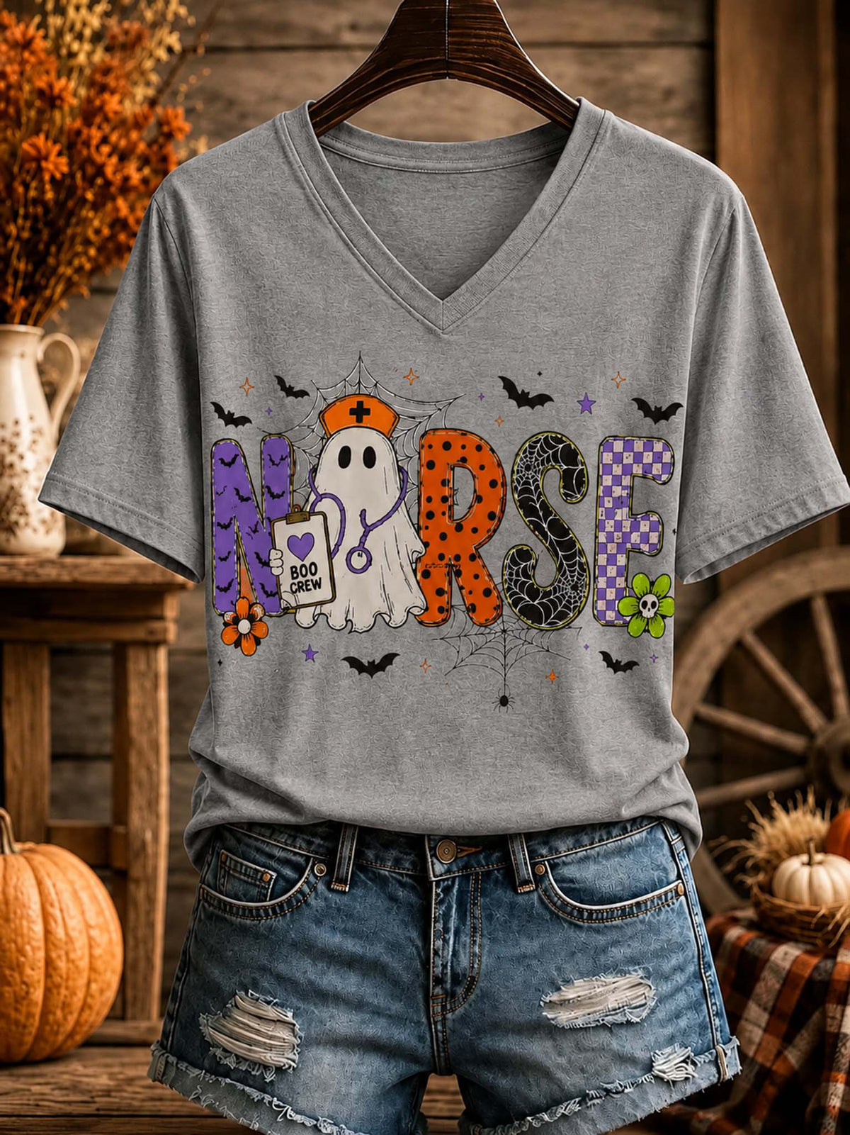 A heather gray V-neck t-shirt with a Halloween-themed 'Nurse' graphic, styled on a wooden hanger with distressed denim shorts against a rustic fall-themed background.