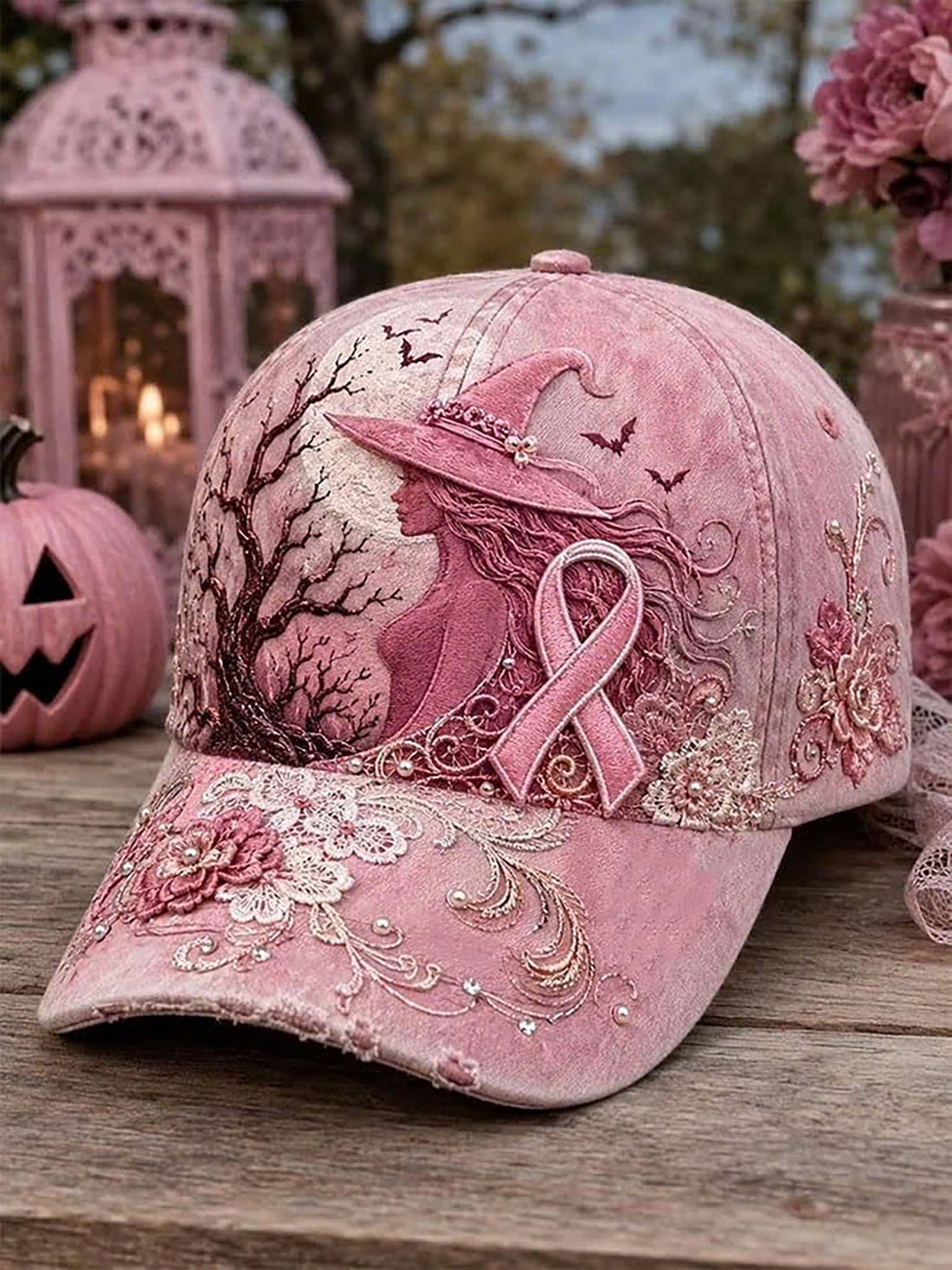 A pink embroidered baseball cap displayed on a wooden surface, featuring a witch silhouette, pink ribbon motif, floral embroidery, and small rhinestone embellishments, with a decorative lantern and flowers in the background.