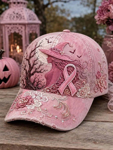 A pink embroidered baseball cap displayed on a wooden surface, featuring a witch silhouette, pink ribbon motif, floral embroidery, and small rhinestone embellishments, with a decorative lantern and flowers in the background.