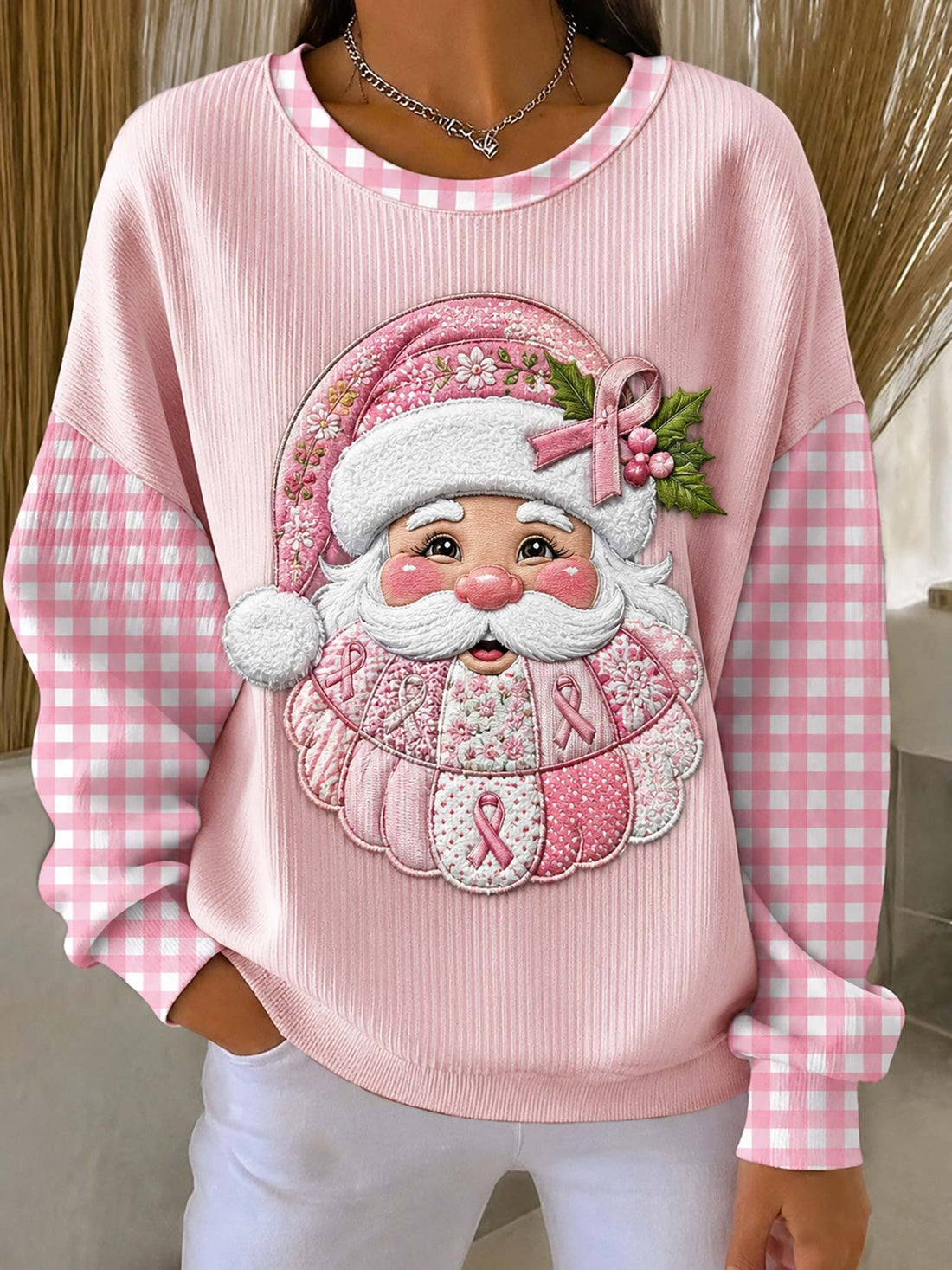 A person wearing a pink ribbed long-sleeve sweatshirt with pink-and-white gingham sleeves and a large embroidered Santa face graphic on the front featuring pink awareness ribbons and green holly leaves.