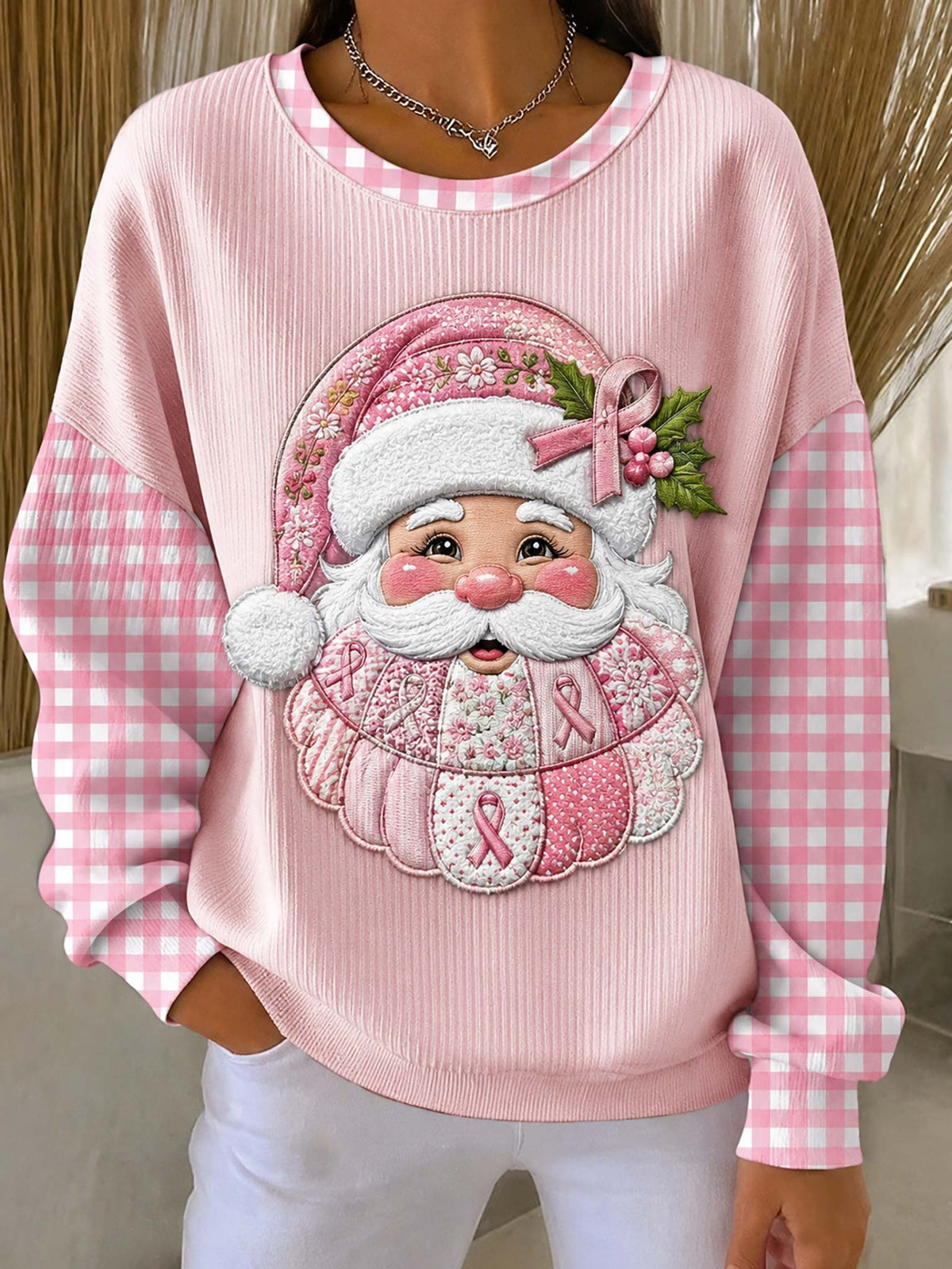 A person wearing a pink ribbed long-sleeve sweatshirt with pink-and-white gingham sleeves and a large embroidered Santa face graphic on the front featuring pink awareness ribbons and green holly leaves.