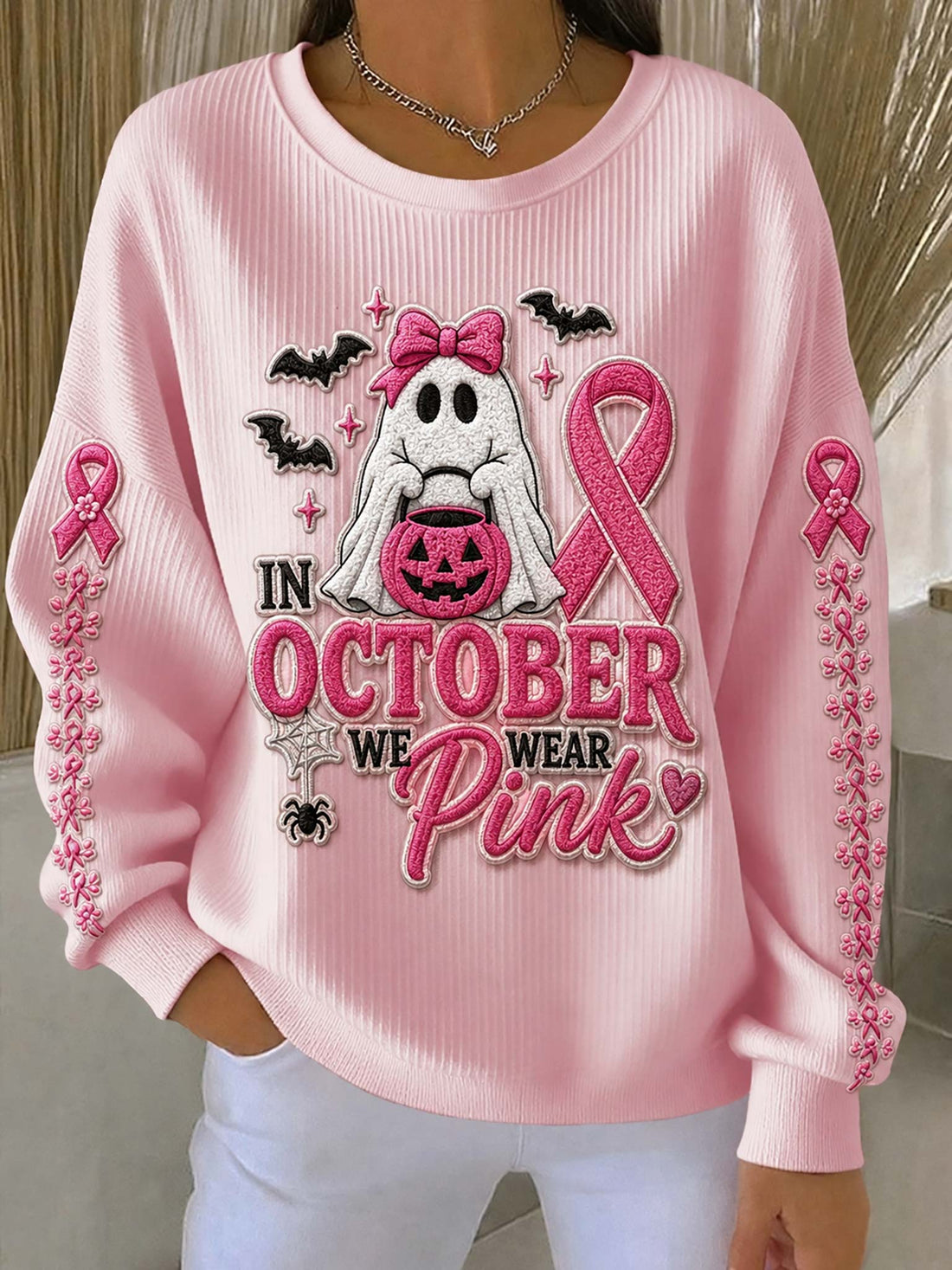 A woman wearing a light pink ribbed long-sleeve sweatshirt with a Halloween-themed graphic that reads 'IN OCTOBER WE WEAR Pink.' The front design includes a white ghost holding a jack-o'-lantern, pink ribbons, bats, and decorative text.