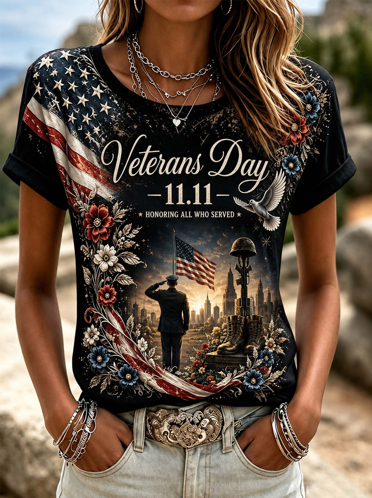 A woman wearing a black graphic t-shirt with a detailed Veterans Day 11.11 design, paired with light-wash jeans and layered silver necklaces and bracelets, set against a blurred outdoor background.
