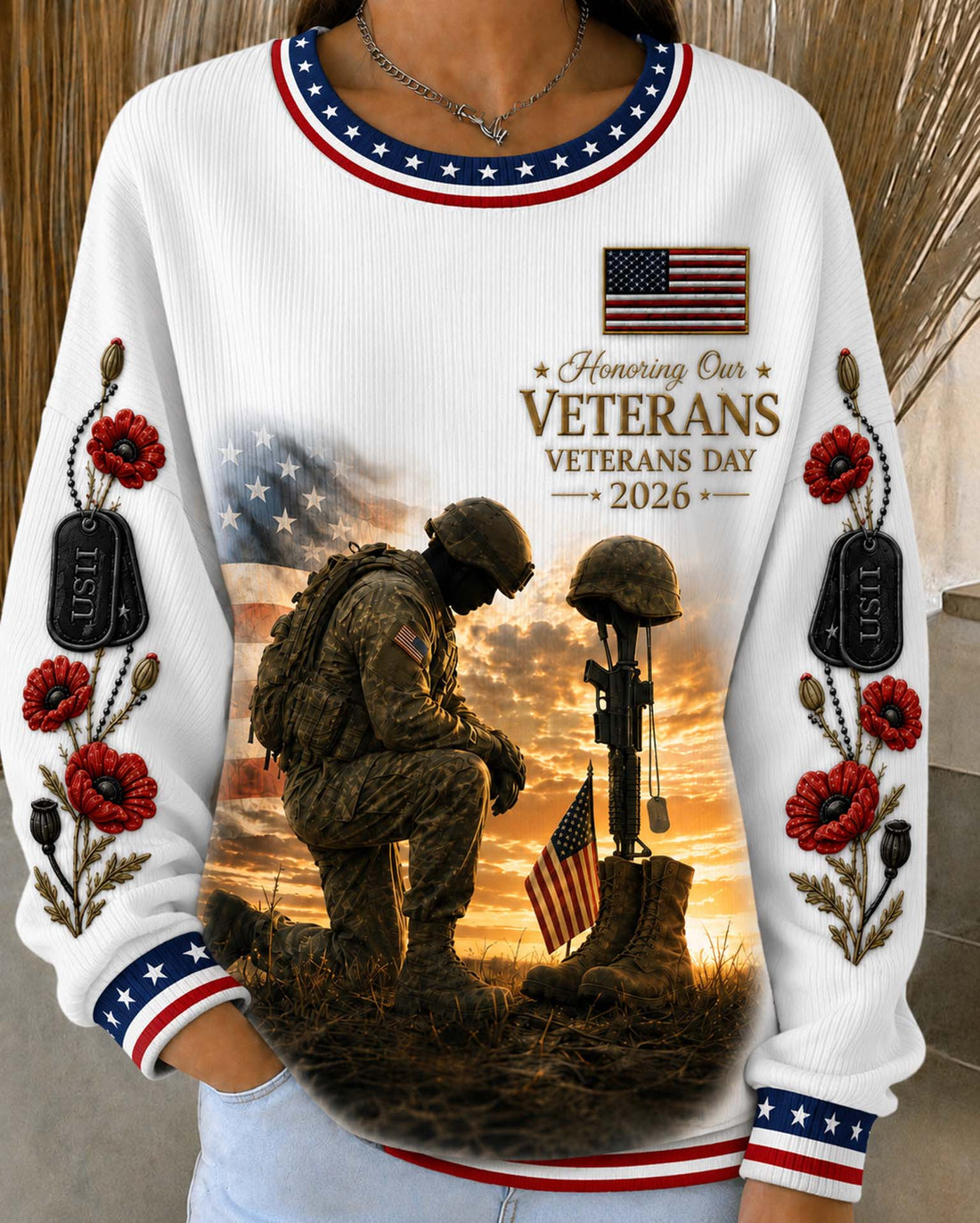 A white long-sleeve crewneck sweatshirt with patriotic red, white, and blue trim, featuring a large graphic print of a kneeling soldier and a helmet on a battlefield at sunset, with 'Veterans Day 2026' text and floral details on the sleeves.
