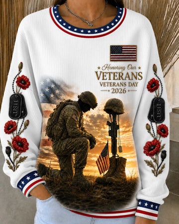 A white long-sleeve crewneck sweatshirt with patriotic red, white, and blue trim, featuring a large graphic print of a kneeling soldier and a helmet on a battlefield at sunset, with 'Veterans Day 2026' text and floral details on the sleeves.
