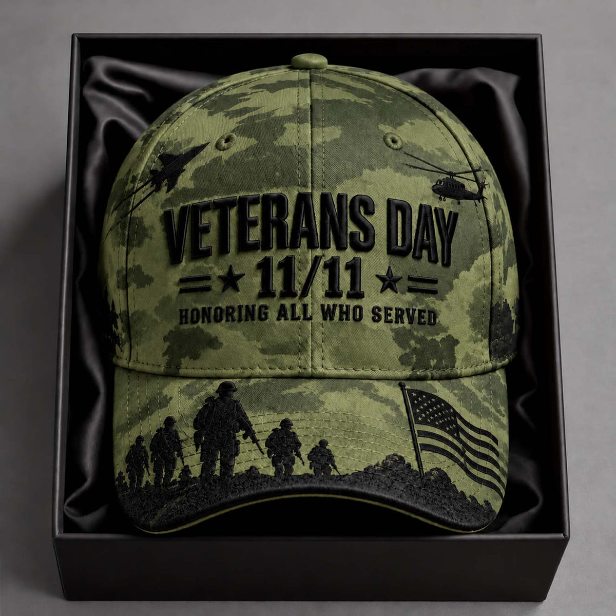 A camouflage baseball cap with black embroidered text reading 'VETERANS DAY 11/11 HONORING ALL WHO SERVED,' plus military silhouettes and an American flag on the brim, presented inside a black gift box.