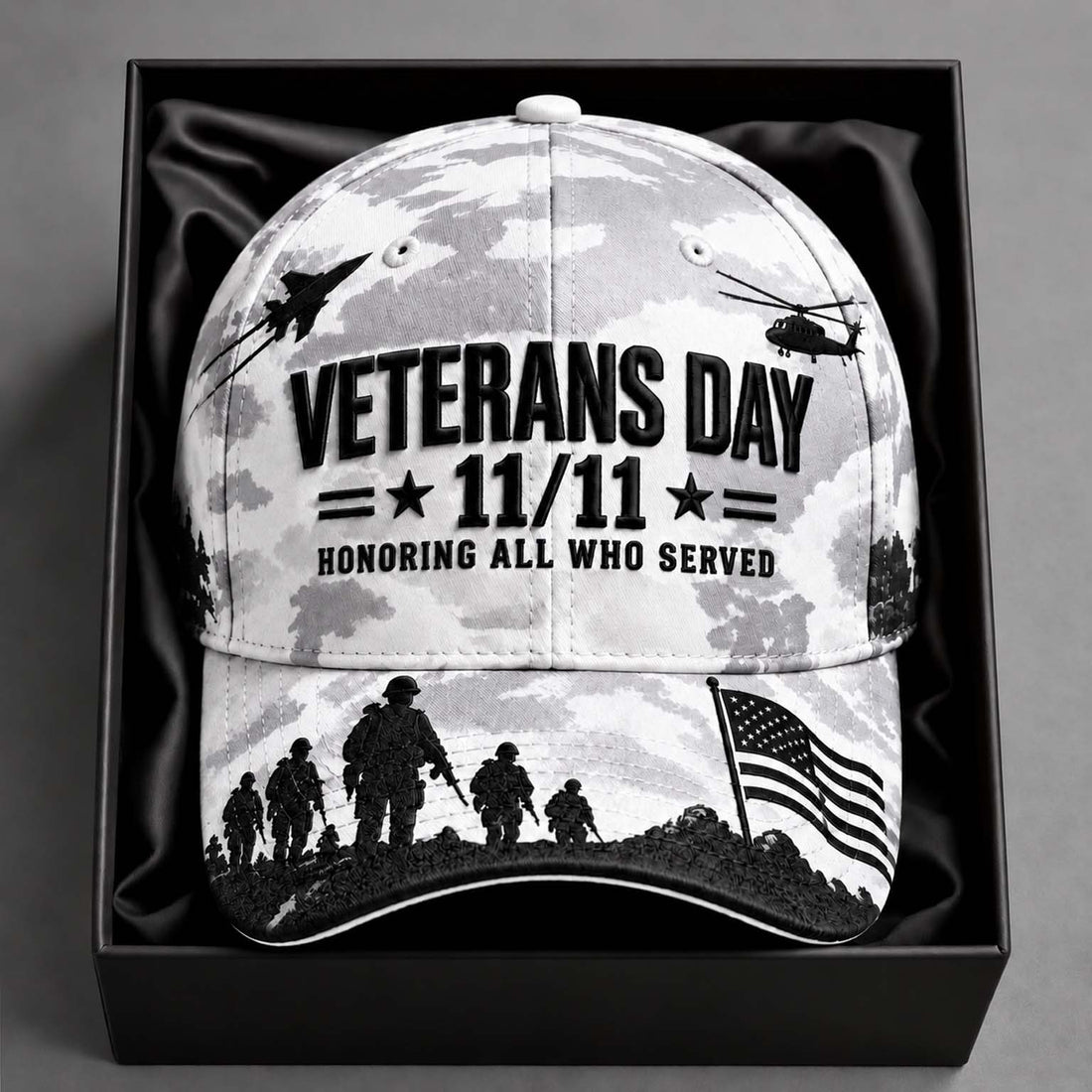 A Veterans Day baseball cap with a black and white camouflage pattern, featuring silhouettes of soldiers, military aircraft, and an American flag, displayed inside a black gift box.
