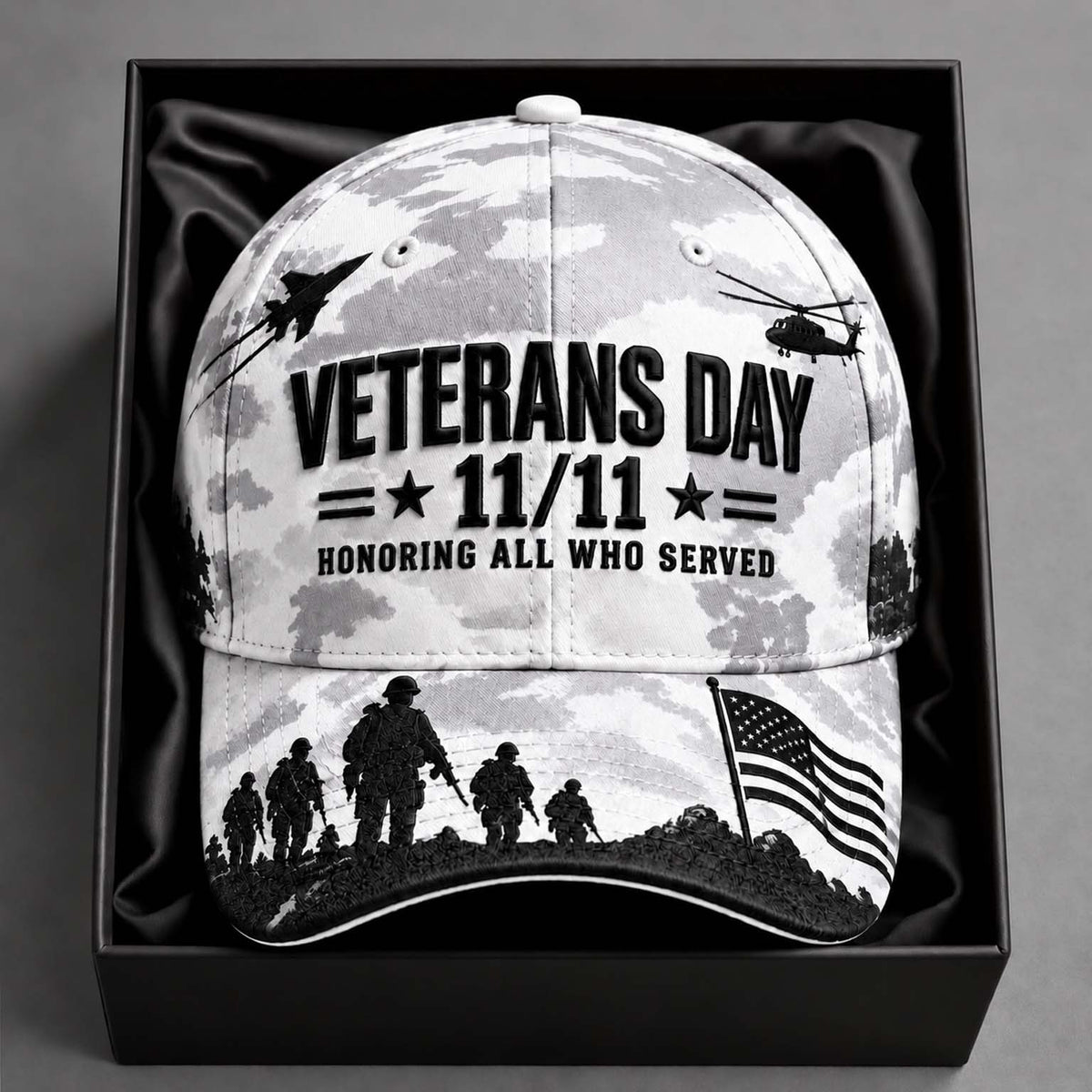 A Veterans Day baseball cap with a black and white camouflage pattern, featuring silhouettes of soldiers, military aircraft, and an American flag, displayed inside a black gift box.