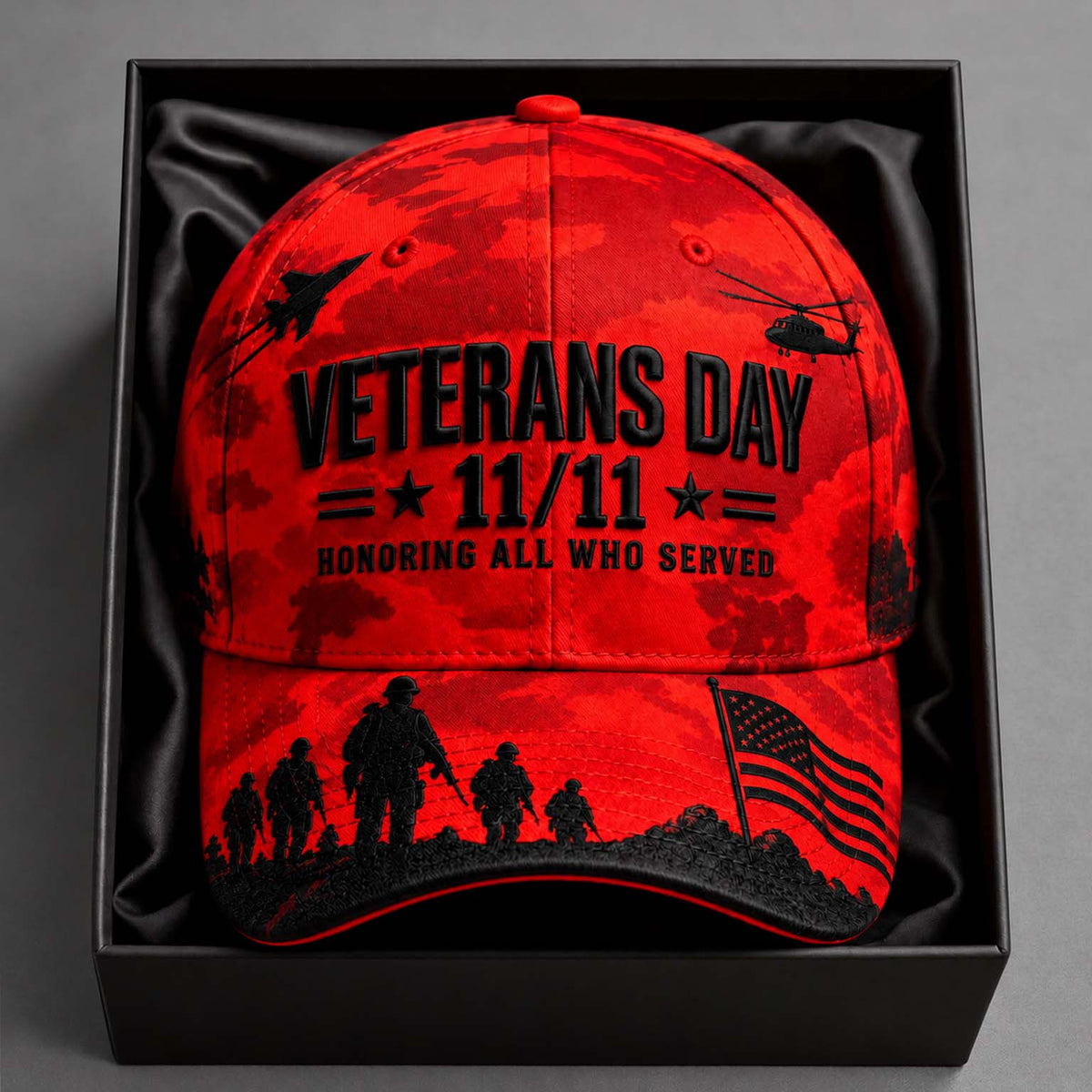 A red camouflage baseball cap with black military-themed graphics and text reading 'Veterans Day 11/11 Honoring All Who Served,' displayed inside a black gift box on a gray background.