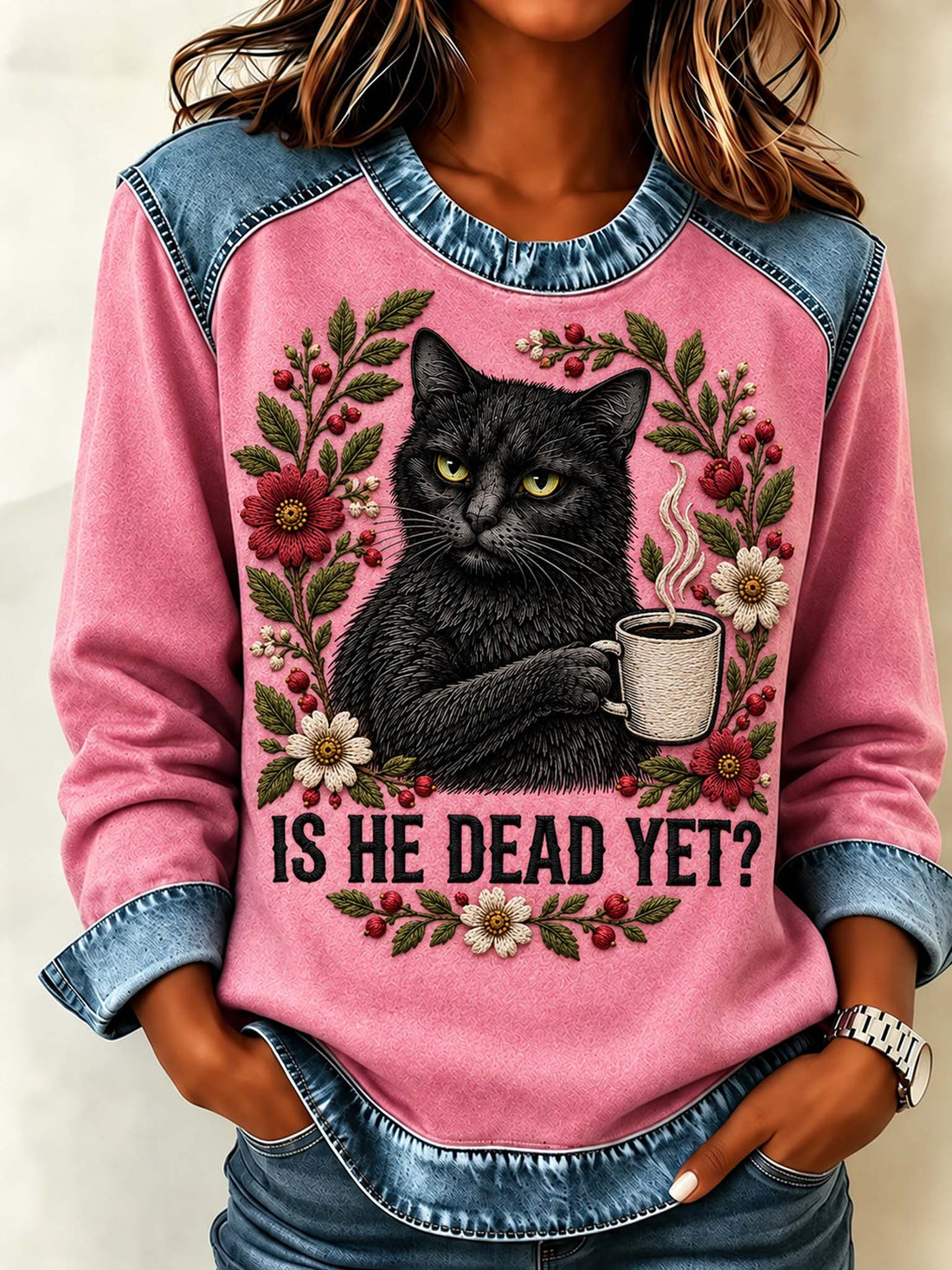 A woman wearing a pink long-sleeve sweatshirt with denim-style shoulder panels and cuffs. The front features a large graphic of a black cat holding a mug, surrounded by floral details and the text 'IS HE DEAD YET?'.