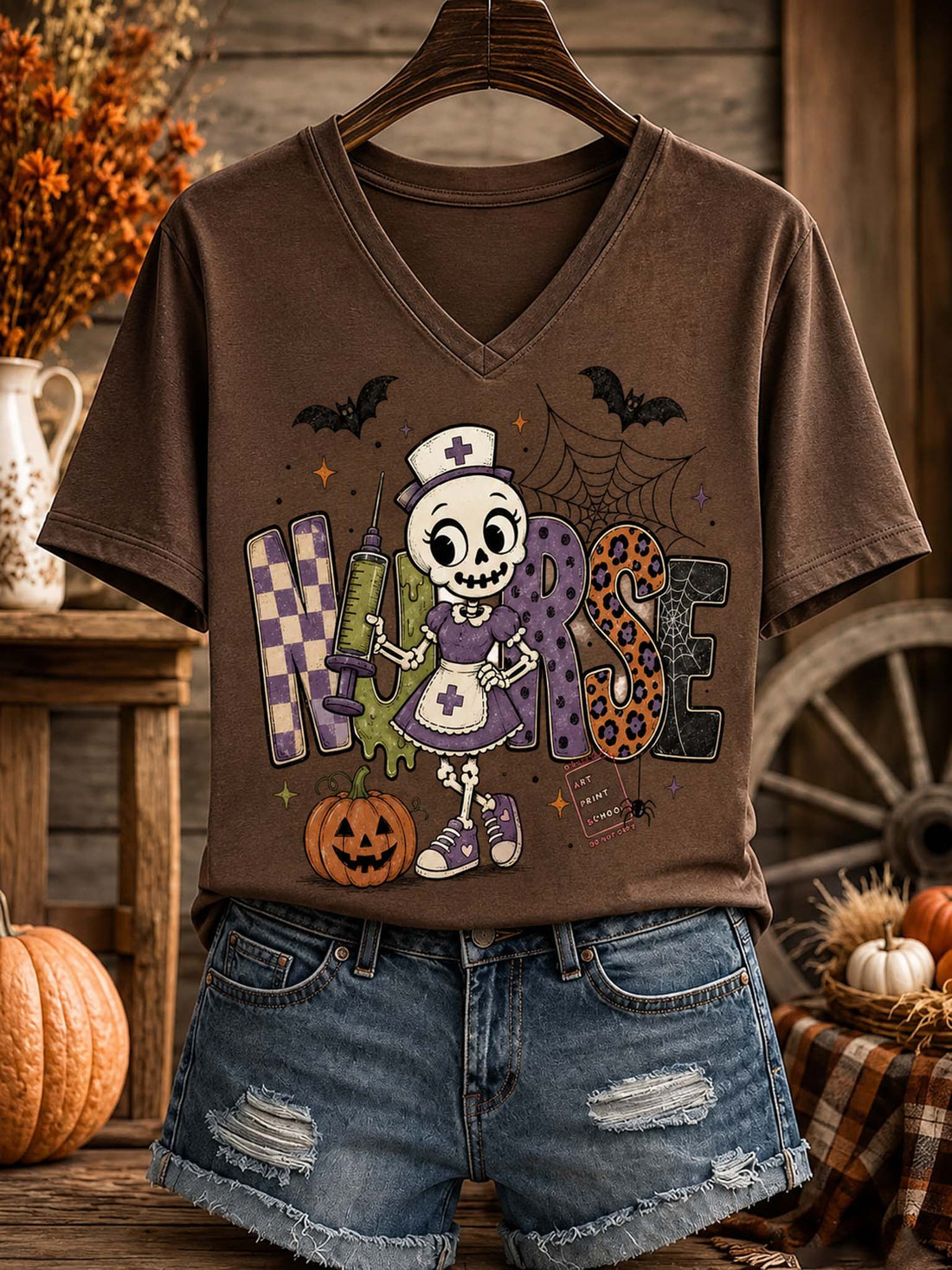 A brown V-neck graphic T-shirt hanging on a wooden hanger, styled with distressed denim shorts in a rustic autumn-themed setting with pumpkins and fall decor.