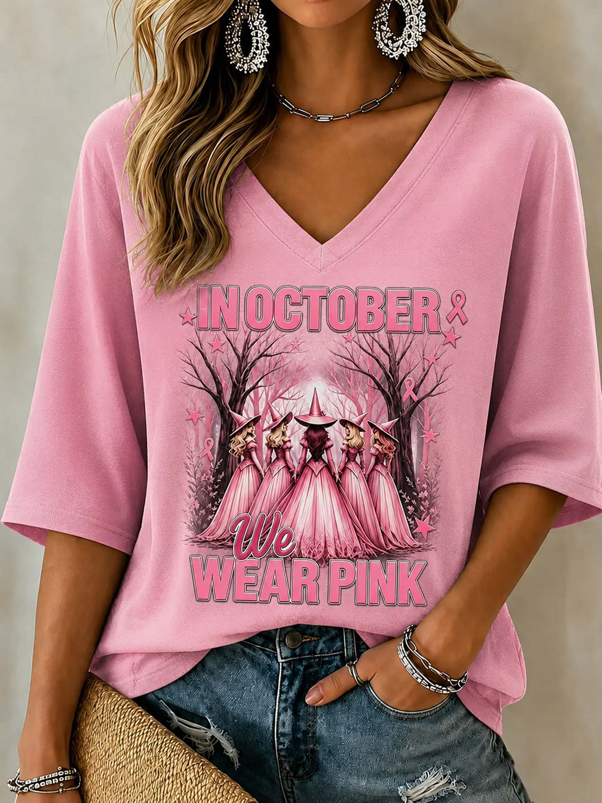 A woman wearing a pink V-neck t-shirt with a 'We Wear Pink' graphic featuring a group of princesses in pink dresses, paired with distressed blue jeans.