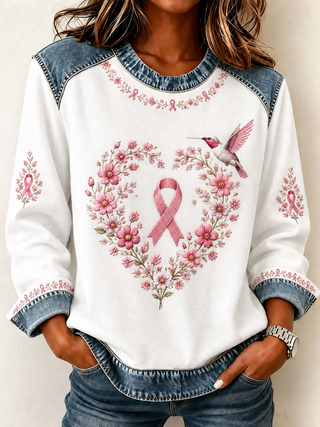 A woman wearing a white long-sleeve sweatshirt with denim shoulder panels and cuffs, featuring a pink breast cancer ribbon surrounded by a floral heart design and a hummingbird on the front.
