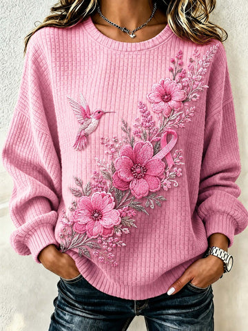 A woman wearing a pink ribbed knit sweater with a large floral and hummingbird embroidery design on the front, paired with dark blue jeans.