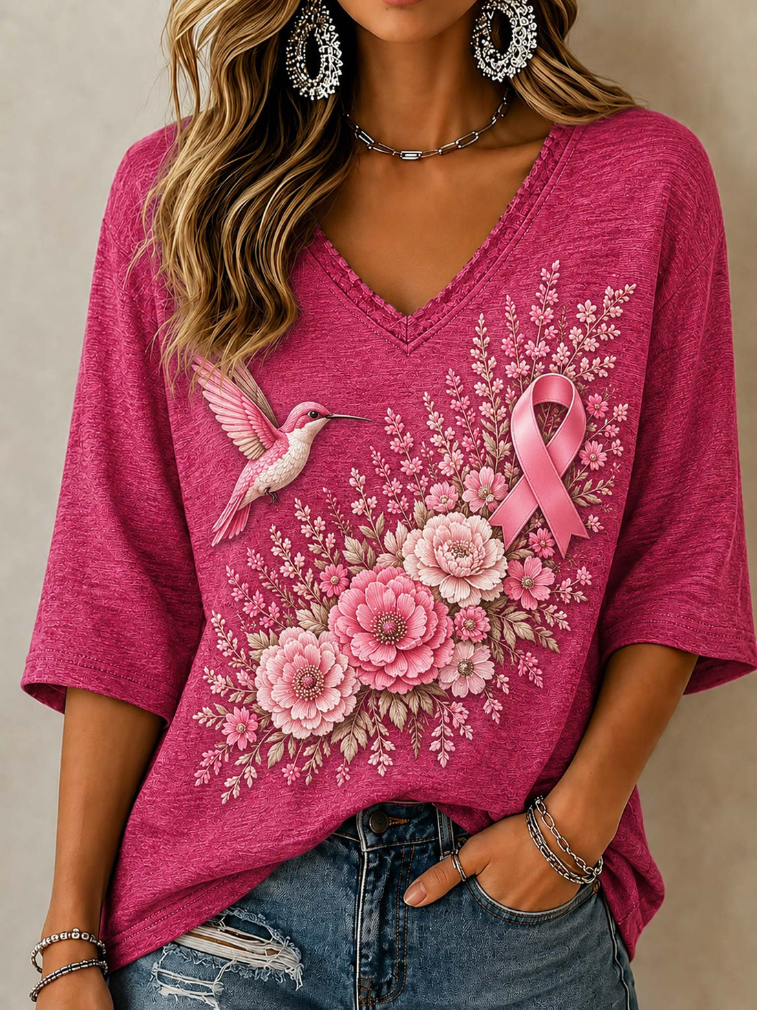 A woman wearing a bright pink V-neck t-shirt with a graphic print of a pink ribbon, a hummingbird, and pink flowers, paired with distressed blue jeans.