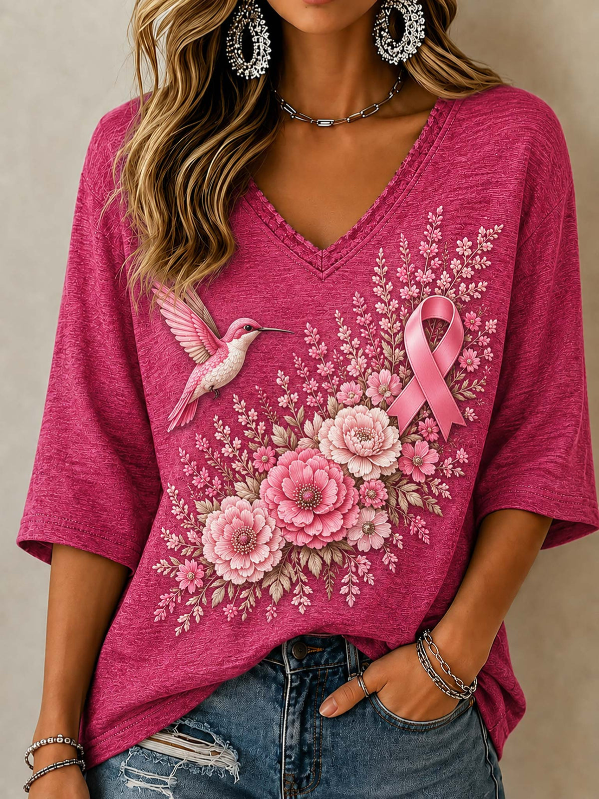 A woman wearing a bright pink V-neck t-shirt with a graphic print of a pink ribbon, a hummingbird, and pink flowers, paired with distressed blue jeans.
