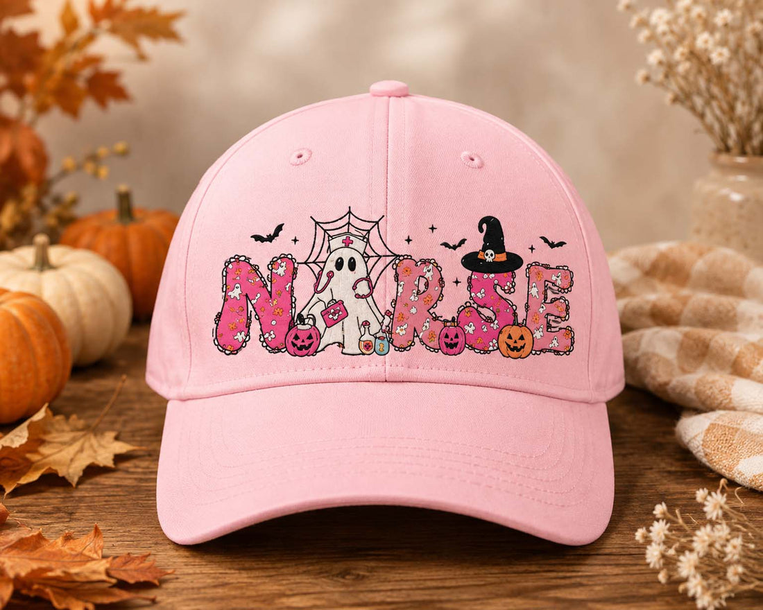 A light pink baseball cap with a 'Nurse' Halloween graphic, displayed on a rustic wooden surface with autumn leaves, pumpkins, and a cozy blanket in the background.