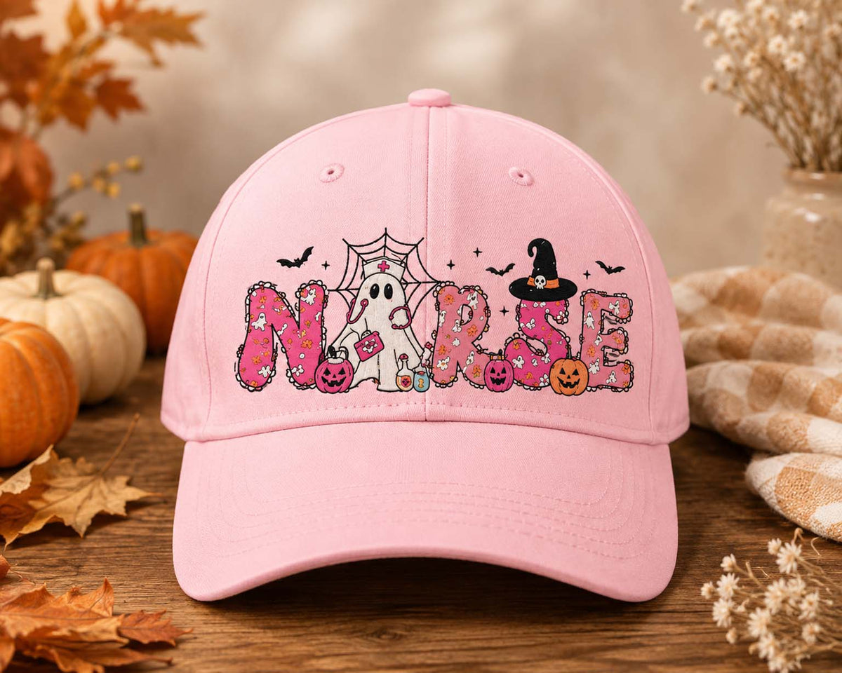 A light pink baseball cap with a 'Nurse' Halloween graphic, displayed on a rustic wooden surface with autumn leaves, pumpkins, and a cozy blanket in the background.