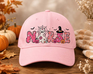 A light pink baseball cap with a 'Nurse' Halloween graphic, displayed on a rustic wooden surface with autumn leaves, pumpkins, and a cozy blanket in the background.