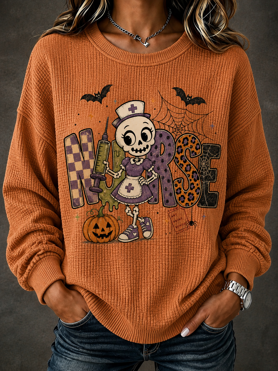 A woman wearing a burnt orange waffle-knit crewneck sweater with a large Halloween nurse graphic on the front, paired with blue jeans against a dark gray background.