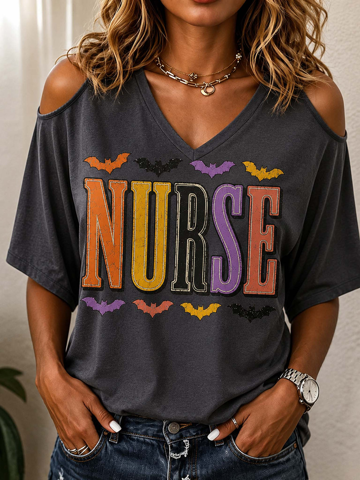 A woman wearing a dark charcoal gray cold-shoulder V-neck t-shirt with a colorful 'NURSE' graphic and bat illustrations, paired with blue jeans.