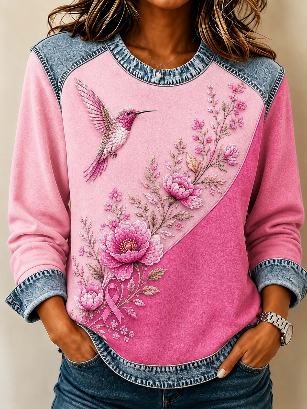 A woman wearing a pink long-sleeve top with a diagonal split design, featuring a large pink flower and hummingbird graphic on one side and a cuffed denim-style sleeve and hem.