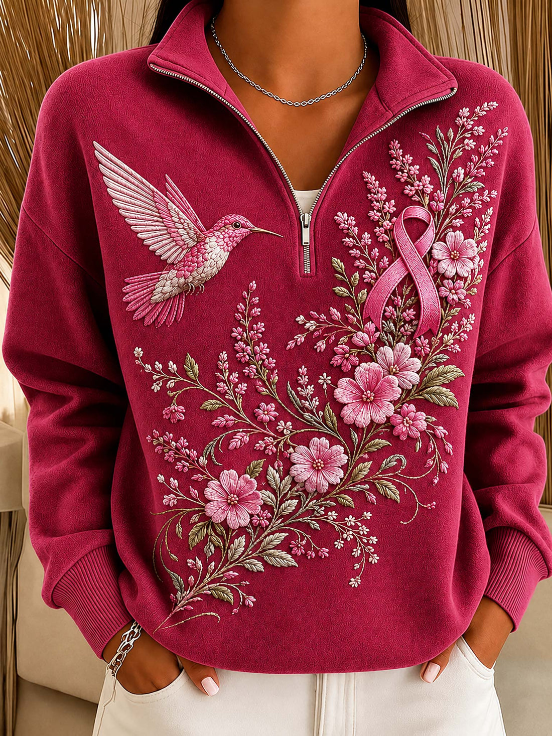 A close-up of a person wearing a magenta pink quarter-zip sweatshirt with a pink ribbon and floral embroidery on the front, paired with white pants.