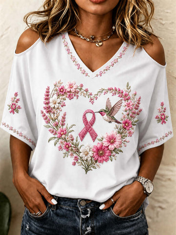 A woman wearing a white cold-shoulder V-neck top with a pink ribbon and floral heart design, paired with dark blue jeans.
