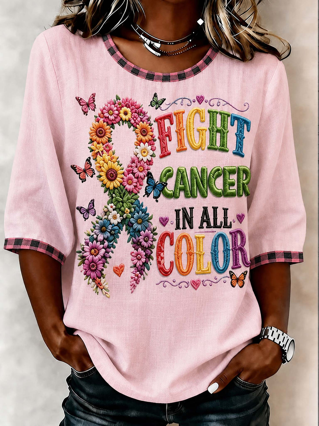 A woman wearing a light pink short-sleeve t-shirt with a colorful graphic that reads 'FIGHT CANCER IN ALL COLOR' alongside a flower and ribbon design, paired with dark jeans.