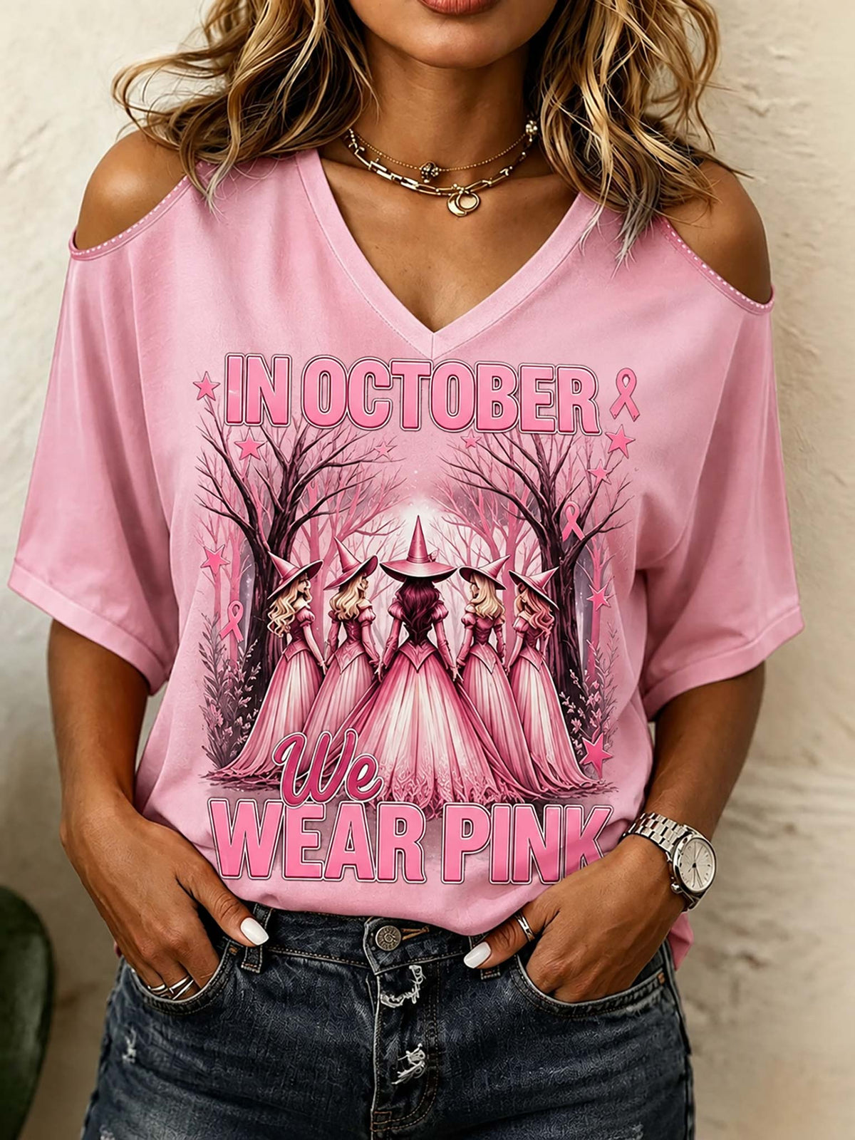 A woman wearing a pink cold-shoulder V-neck t-shirt with a graphic of five witches in a forest and the text 'IN OCTOBER WE WEAR PINK'.