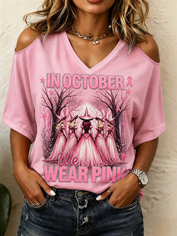 A woman wearing a pink cold-shoulder V-neck t-shirt with a graphic of five witches in a forest and the text 'IN OCTOBER WE WEAR PINK'.