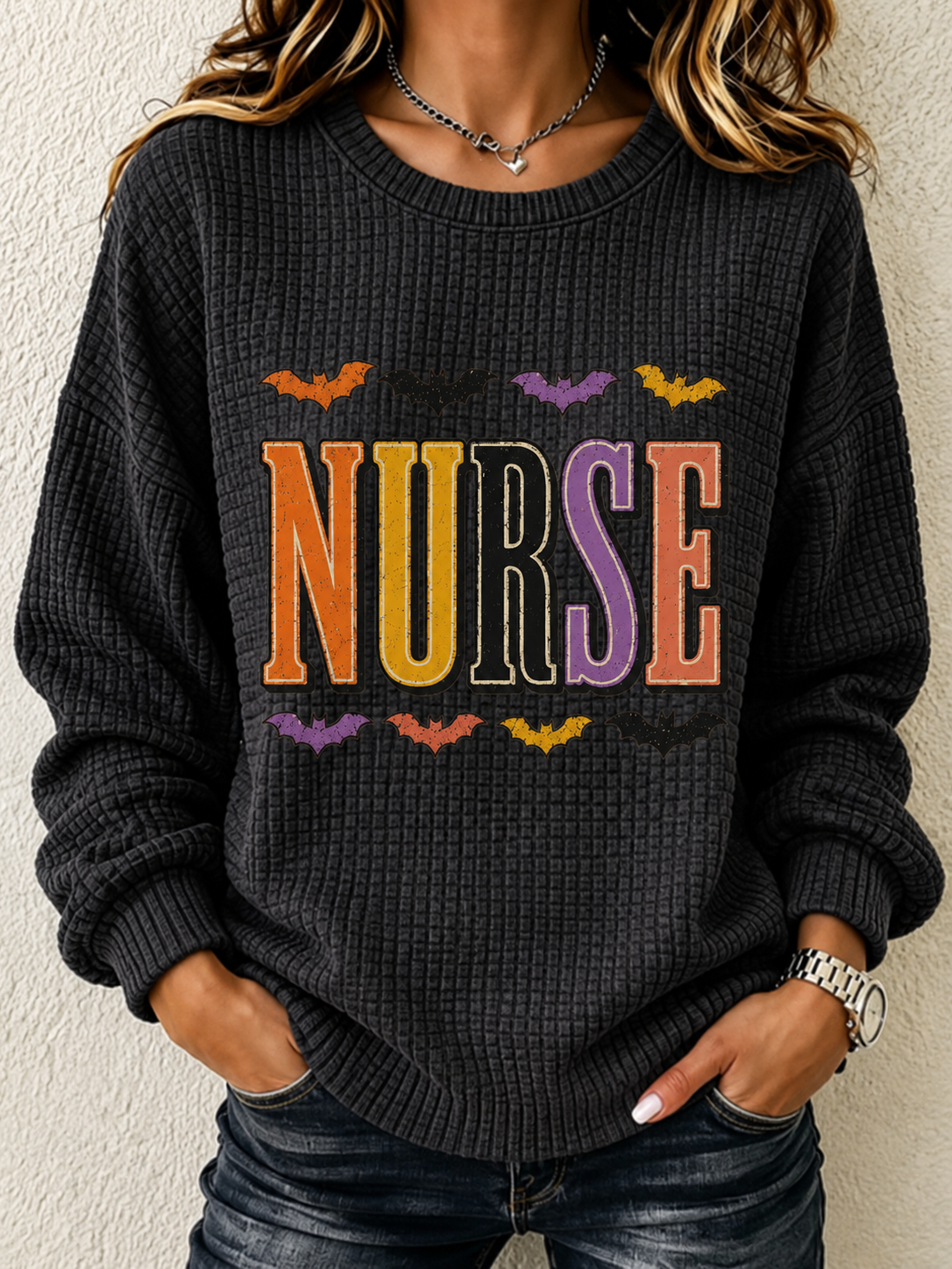 A woman wearing a dark charcoal gray waffle-knit sweater with a colorful 'NURSE' graphic and bat motifs, paired with dark blue jeans.