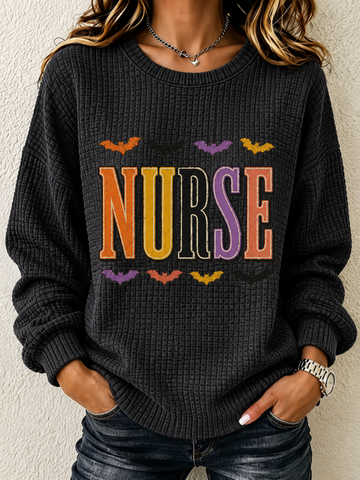 A woman wearing a dark charcoal gray waffle-knit sweater with a colorful 'NURSE' graphic and bat motifs, paired with dark blue jeans.