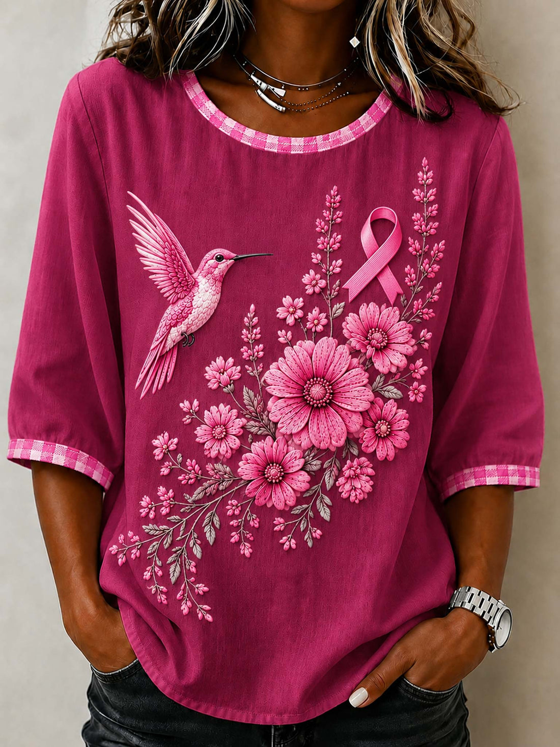 A woman wearing a magenta pink short-sleeve top with a pink and white gingham trim, featuring a graphic print of a pink hummingbird, pink flowers, and a pink awareness ribbon.