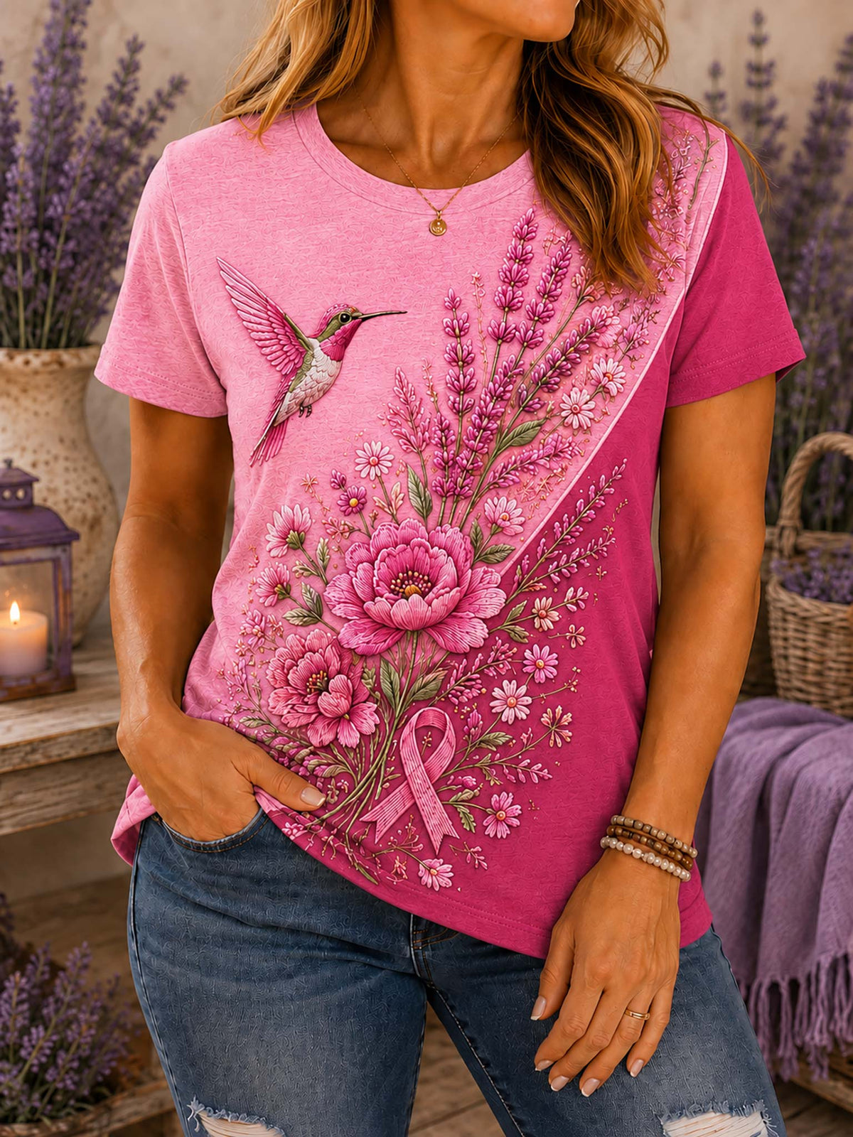 A woman wearing a pink graphic t-shirt with a floral print of a hummingbird, flowers, and a pink ribbon, paired with blue jeans in a lavender-themed setting.