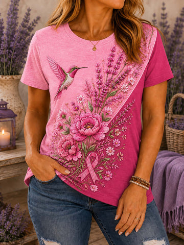 A woman wearing a pink graphic t-shirt with a floral print of a hummingbird, flowers, and a pink ribbon, paired with blue jeans in a lavender-themed setting.