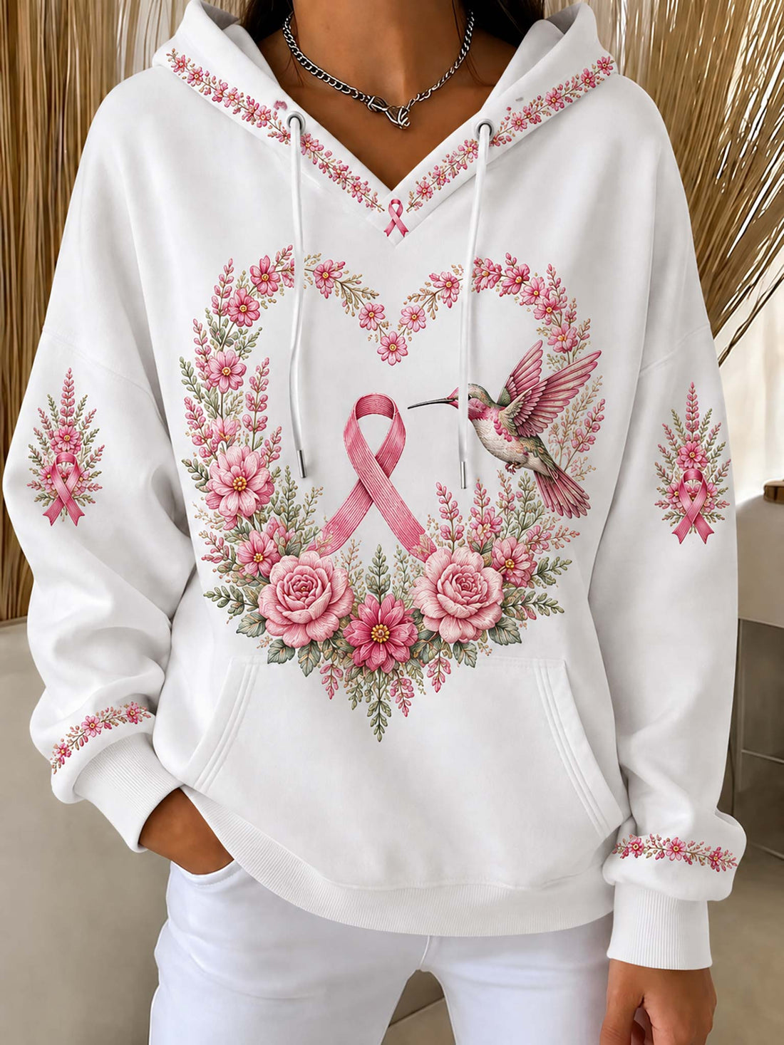 A white pullover hoodie with a pink breast cancer awareness ribbon, a pink and green floral wreath, and a hummingbird graphic on the front. The hoodie features pink floral embroidery details on the hood, cuffs, and sleeves, and is modeled with white pants.