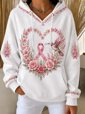 A white pullover hoodie with a pink breast cancer awareness ribbon, a pink and green floral wreath, and a hummingbird graphic on the front. The hoodie features pink floral embroidery details on the hood, cuffs, and sleeves, and is modeled with white pants.