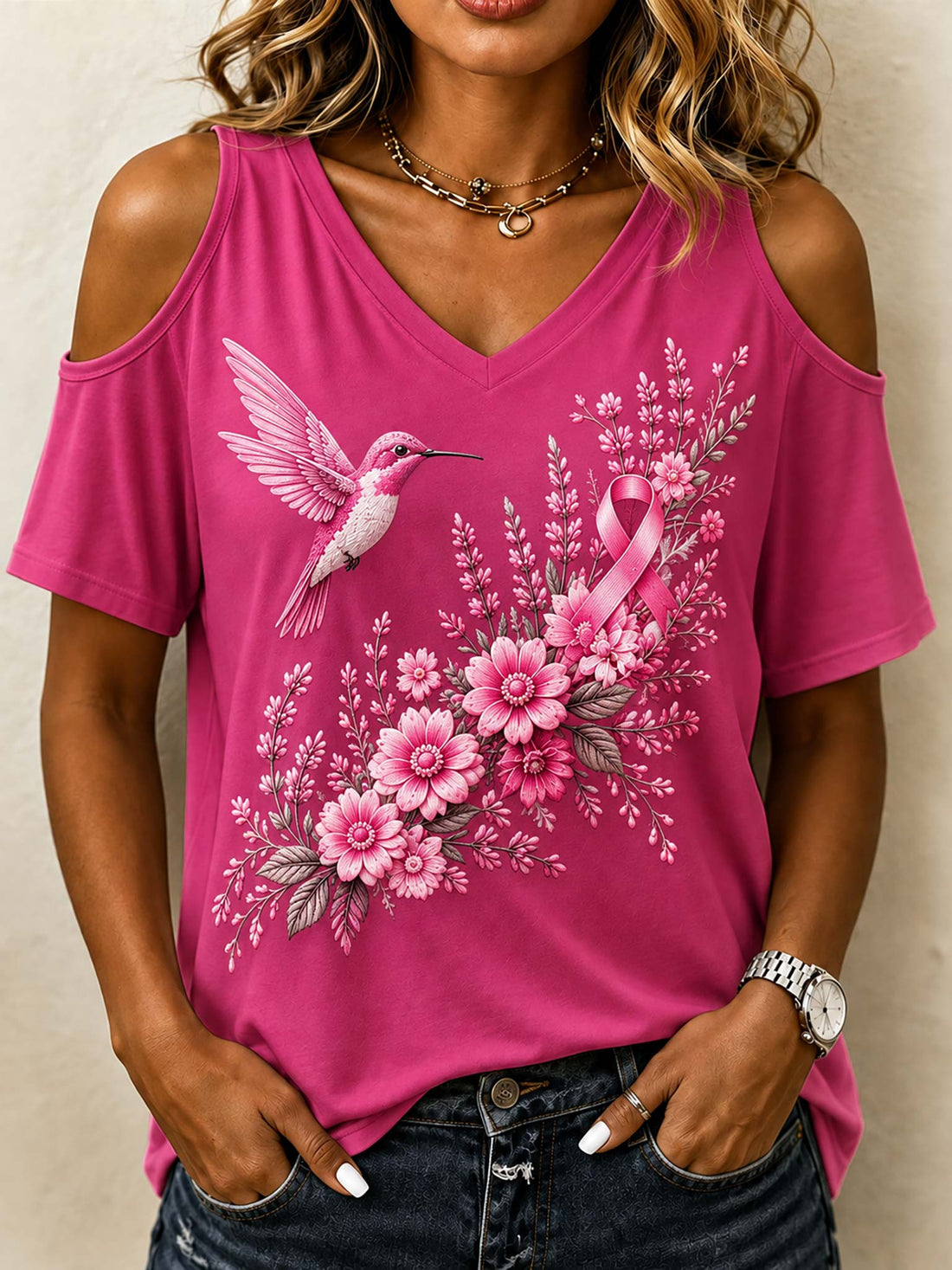 A woman wearing a bright pink cold-shoulder V-neck top with a graphic print of a pink hummingbird, pink ribbon, and pink flowers, paired with dark blue jeans.