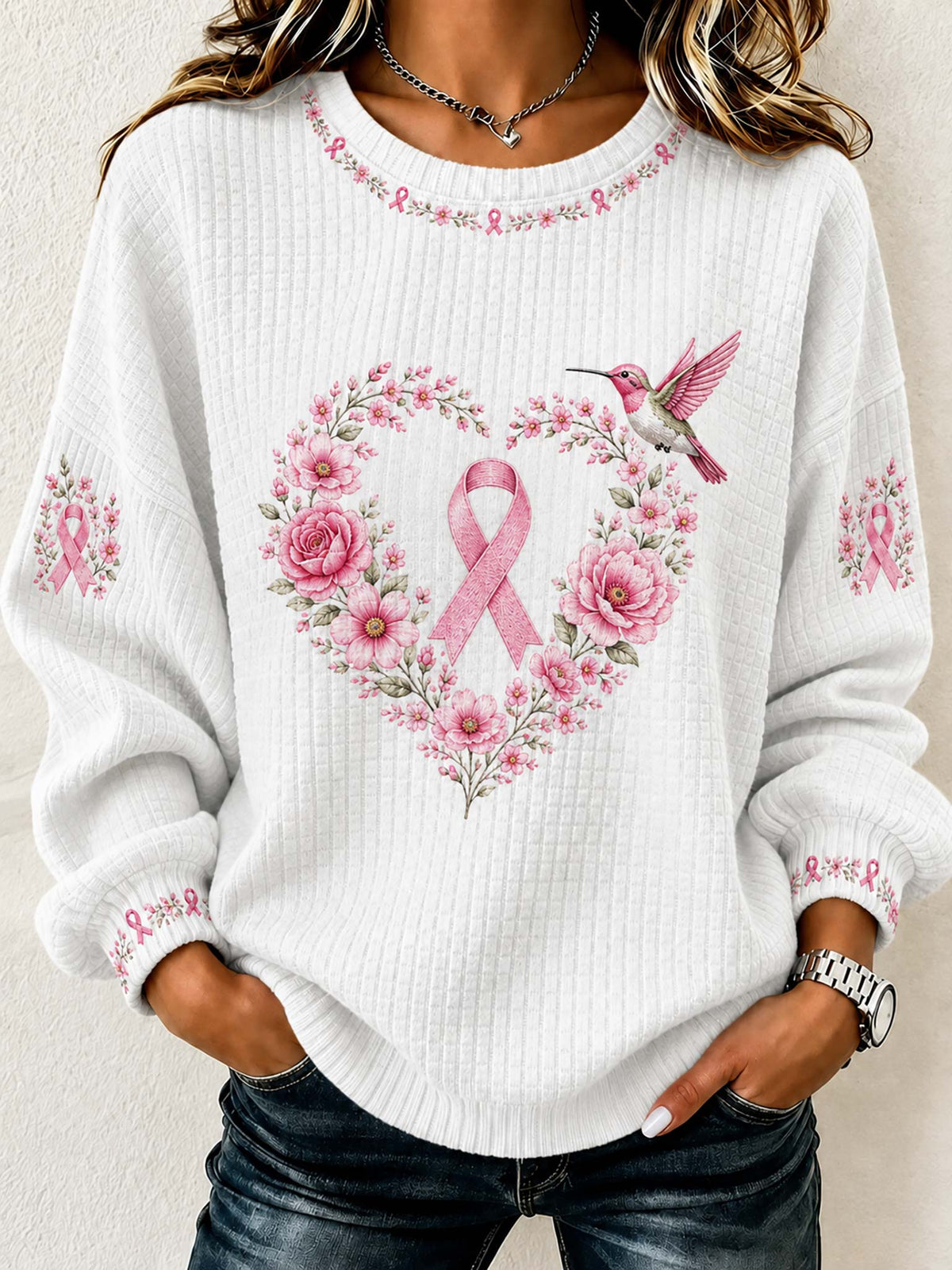 A woman wearing a white long-sleeve waffle-knit sweater with a pink floral heart design, pink ribbon, and hummingbird on the front, paired with dark blue jeans.