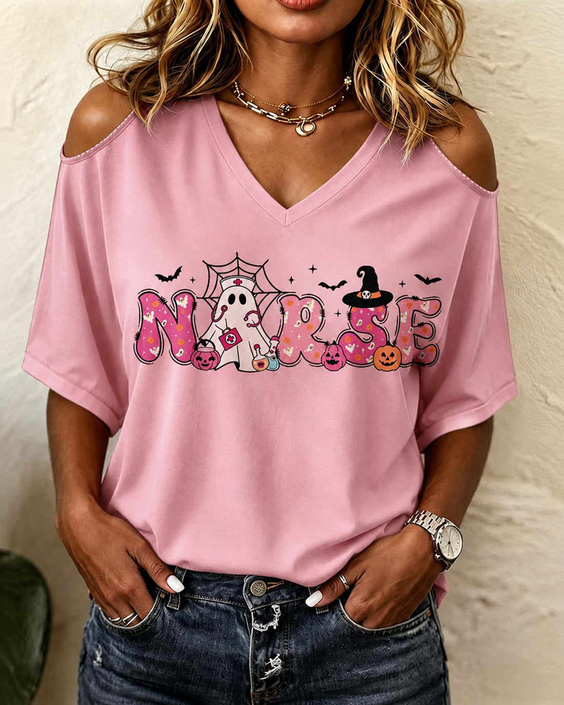 A woman wearing a pink cold-shoulder V-neck t-shirt with a 'NORSE' Halloween graphic, paired with distressed blue jeans.